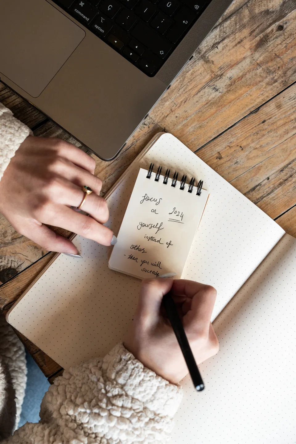 Hands writing personal notes and goals in a notebook beside a laptop